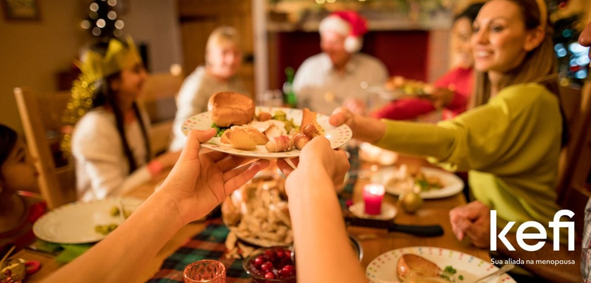 A menopausa pode atrapalhas as festas de final de ano. A imagem representa uma família reunida durante as festas de fim de ano compartilhando uma refeição saudável à mesa, com destaque para a troca de pratos e o ambiente decorado com tema natalino. Na lateral inferior direita a logo da marca Kefi, sua aliada na menopausa.