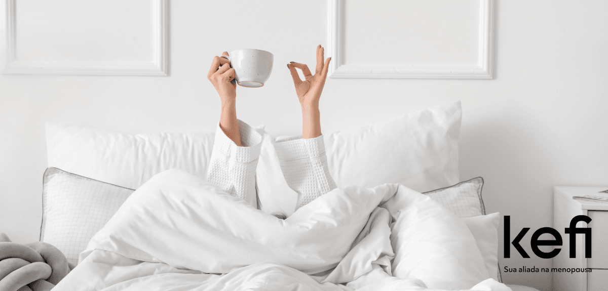 Mulher relaxando em uma cama confortável, com as mãos levantadas segurando uma xícara de chá, simbolizando calma e inteligência emocional na menopausa.