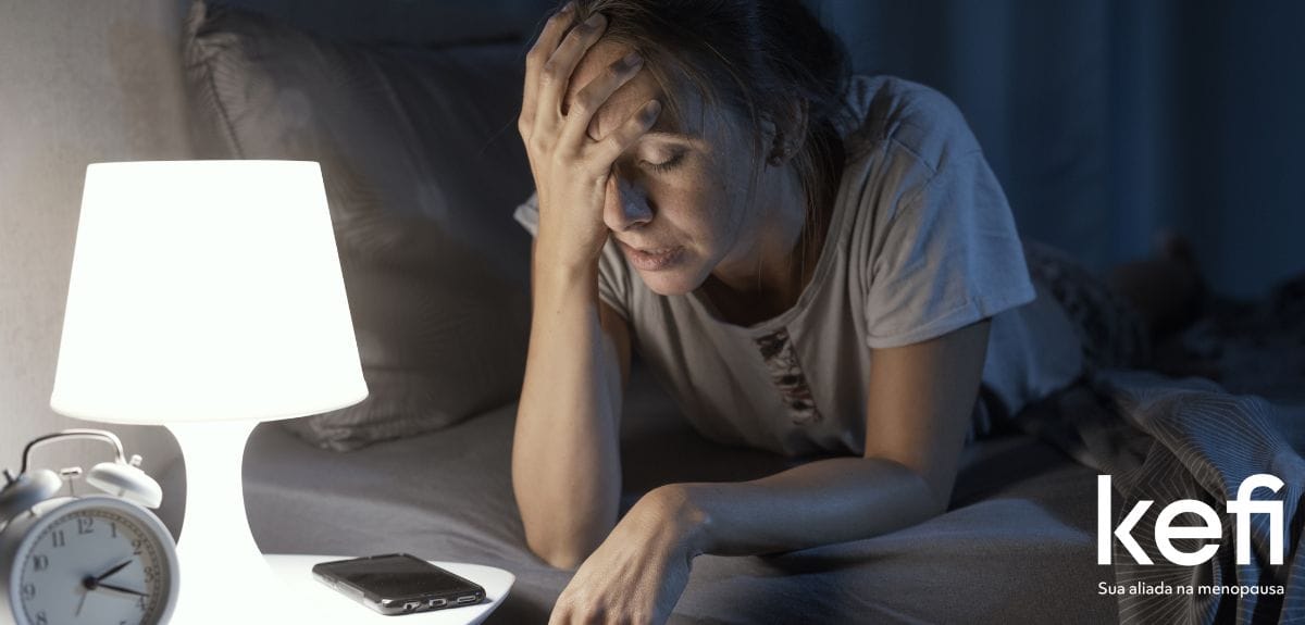 Mulher deitada na cama, com expressão de cansaço e preocupação, segurando a cabeça enquanto encara o celular e o relógio, representando a insônia na menopausa.