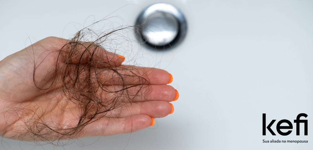 Mão feminina segurando grande quantidade de fios de cabelo caídos sobre a pia, representando a queda de cabelo na menopausa.